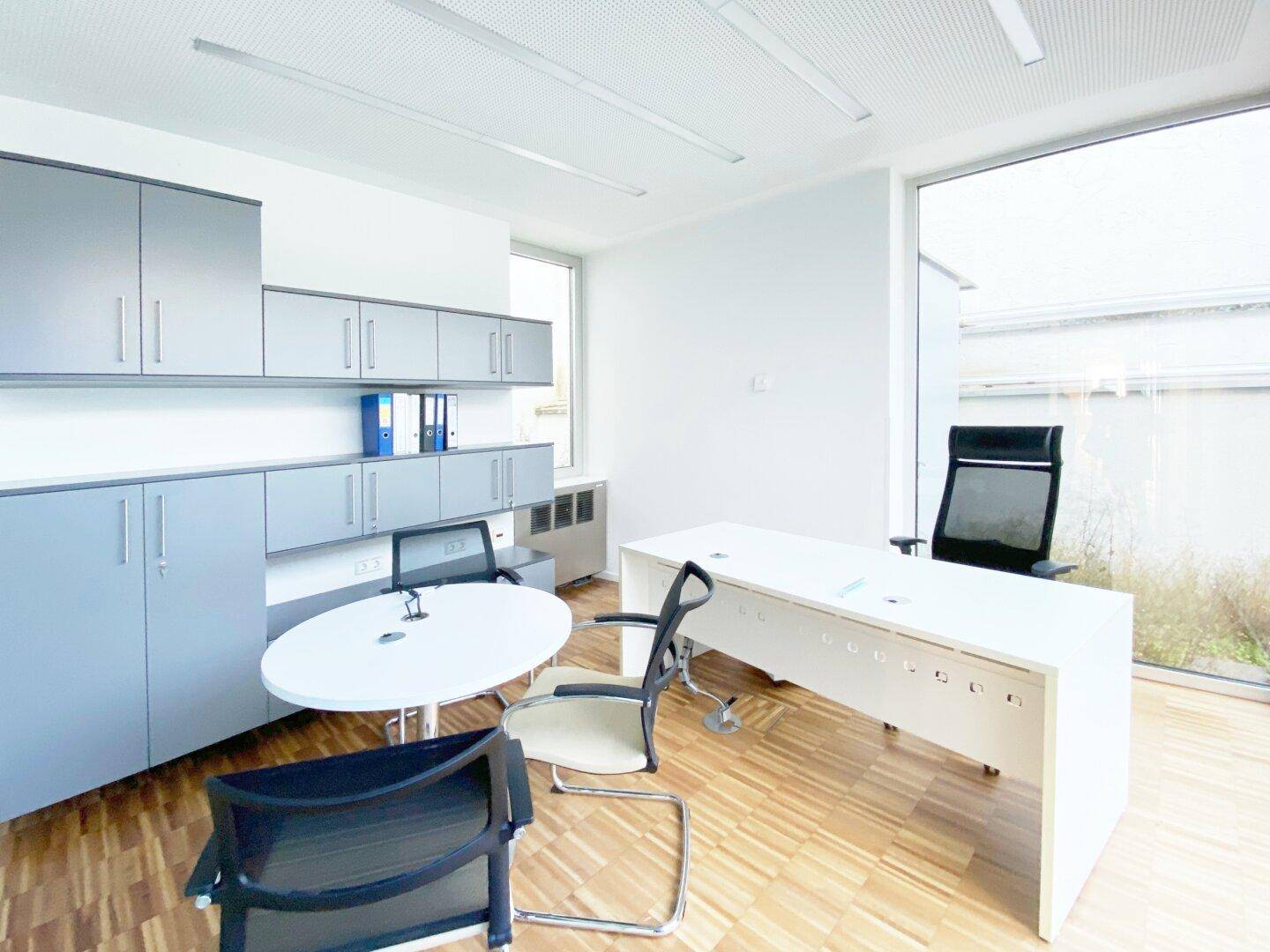 Geräumiges Büro mit umfangreichen grauen Einbauschränken, einem großen Schreibtisch und einem Fenster mit Blick ins Grüne.