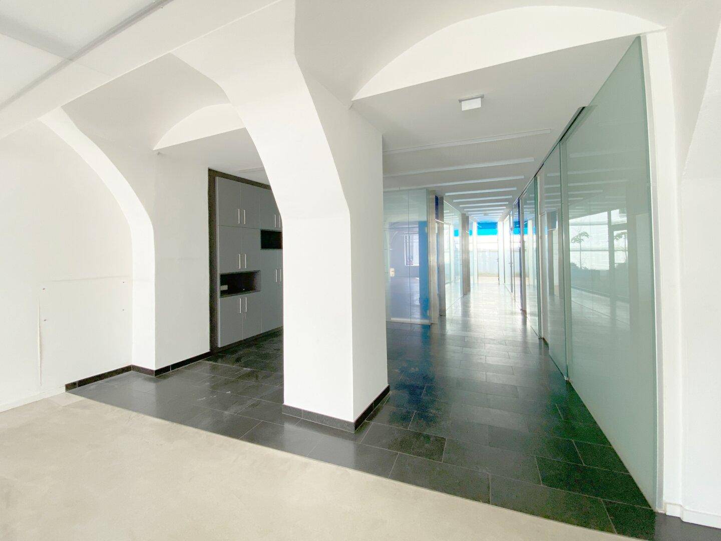 Flurartiger Bereich im Gewerberaum mit dunklen Fliesen, Säulen und modernen Glasabtrennungen für Büros.