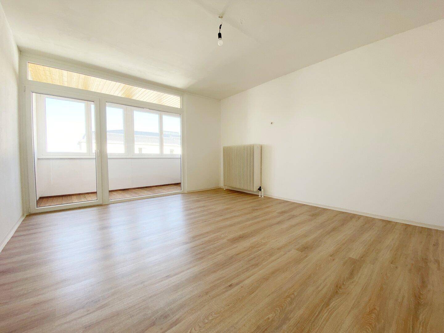 Geräumiges Wohnzimmer mit hellem Laminatboden und viel Tageslicht durch die großen Fenster zur Loggia.