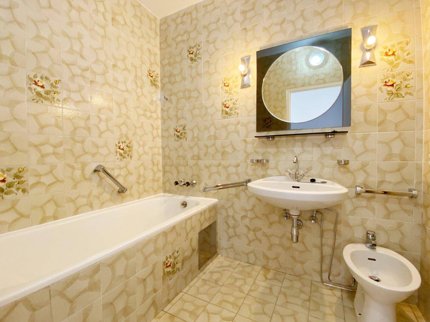 Badezimmer im 80er-Jahre-Stil mit beigefarbenen Fliesen, Badewanne, Waschbecken, Bidet und Haltegriffen.
