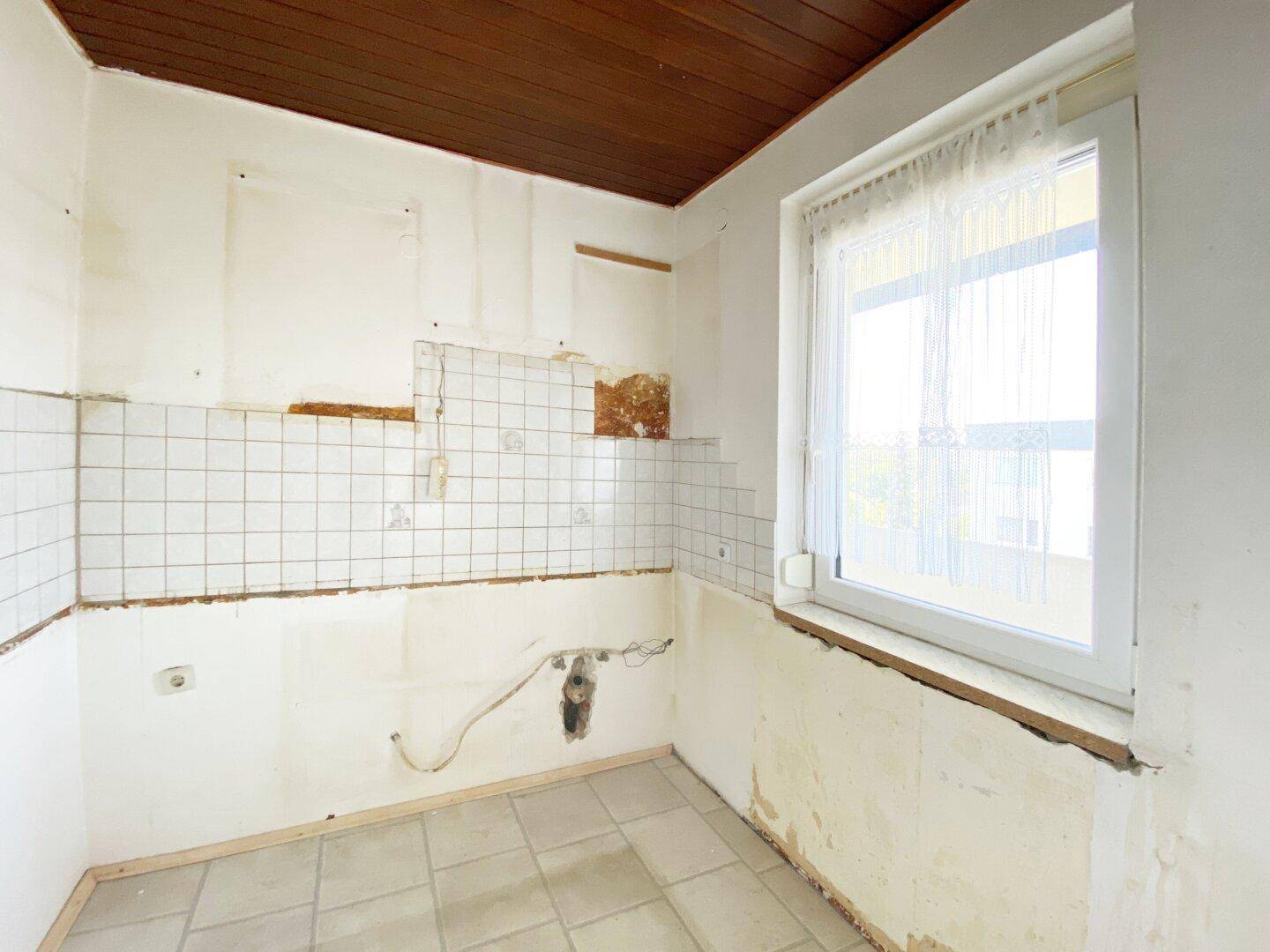 Leere Küche mit Fenster, teilweise entfernten Fliesen und Holzdeckenverkleidung, bereit zur Neugestaltung.
