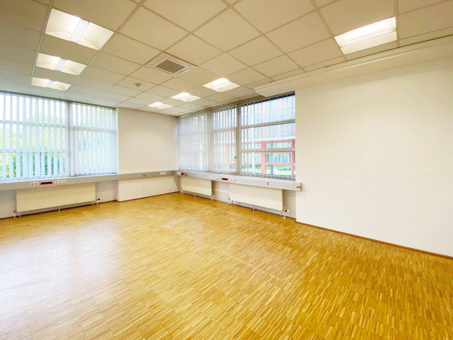 Helles Büro mit Parkettboden und großen Fenstern, die mit Jalousien ausgestattet sind.