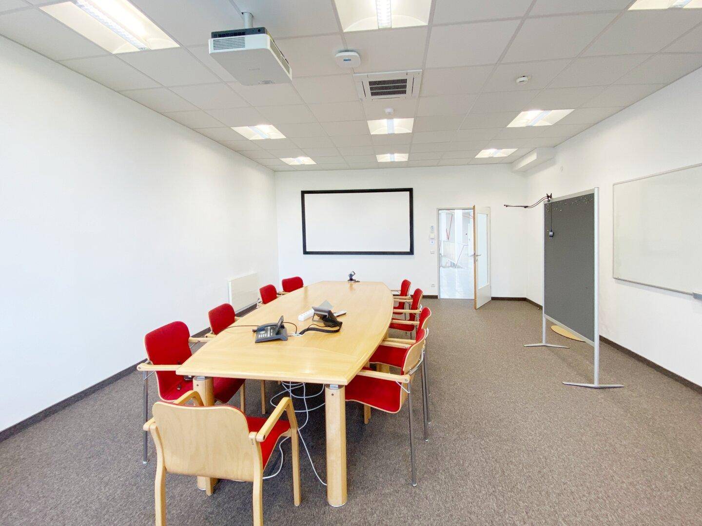 Konferenzraum mit Beamer, Leinwand und großem Holztisch für Besprechungen.