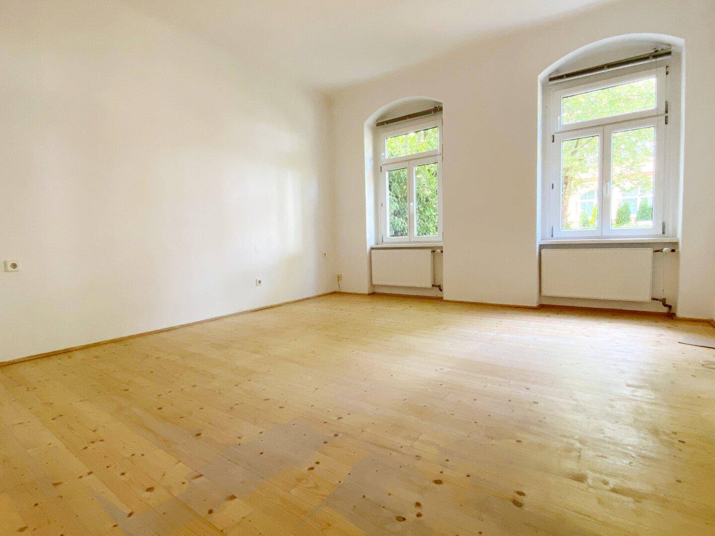 Leerer Raum mit Holzboden, weißen Wänden und zwei großen Fenstern.