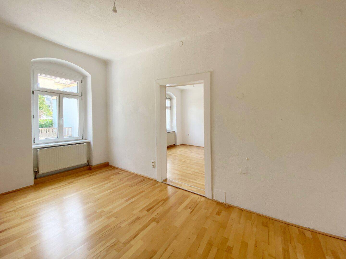 Heller Raum mit Holzboden, weißen Wänden und einem Durchgang zu einem weiteren Zimmer.