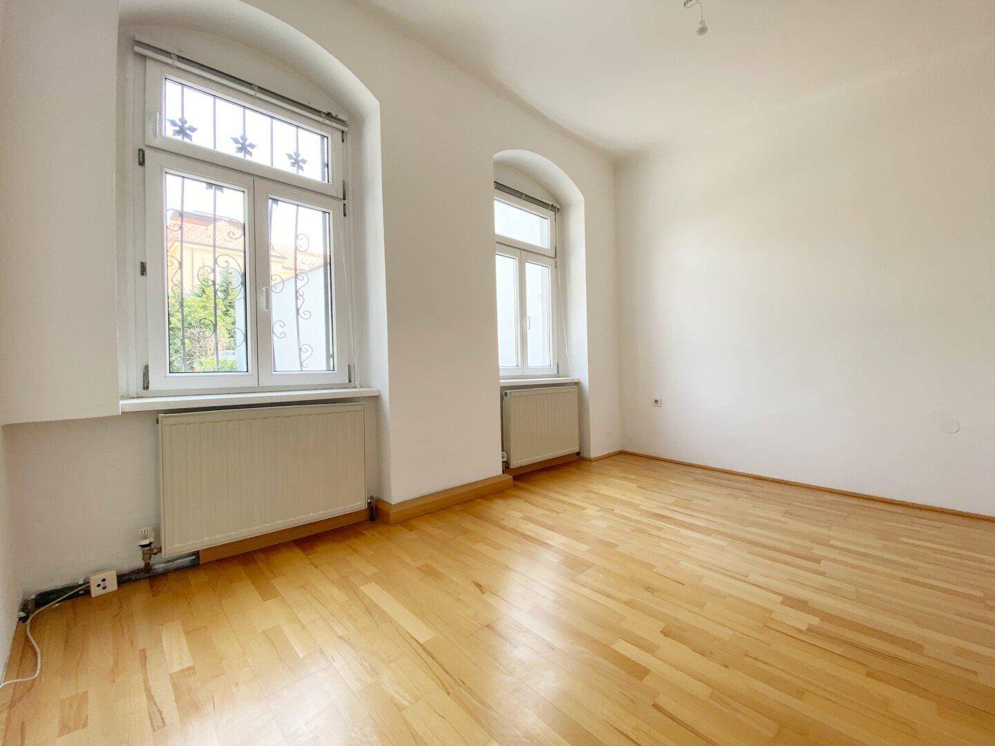 Geräumiger Raum mit Holzboden, weißen Wänden und zwei Fenstern mit Gitter.