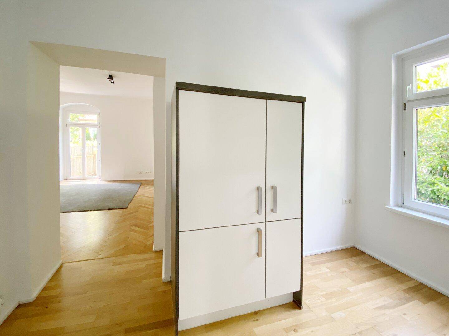 Heller Raum mit Parkettboden und einem Schrank, der zu einem weiteren Wohnbereich führt.
