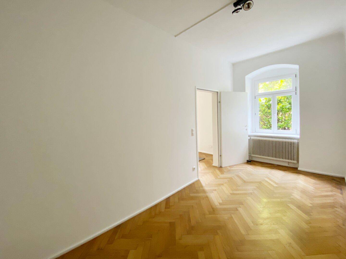 Geräumiger Raum mit klassischem Fischgrätparkett, großem Fenster und Heizkörper unter dem Fenster.