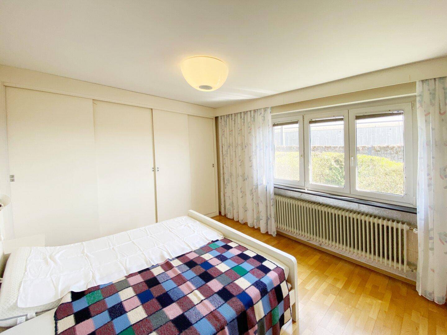 Helles Schlafzimmer mit großem Fenster, Heizkörper und Parkettboden.