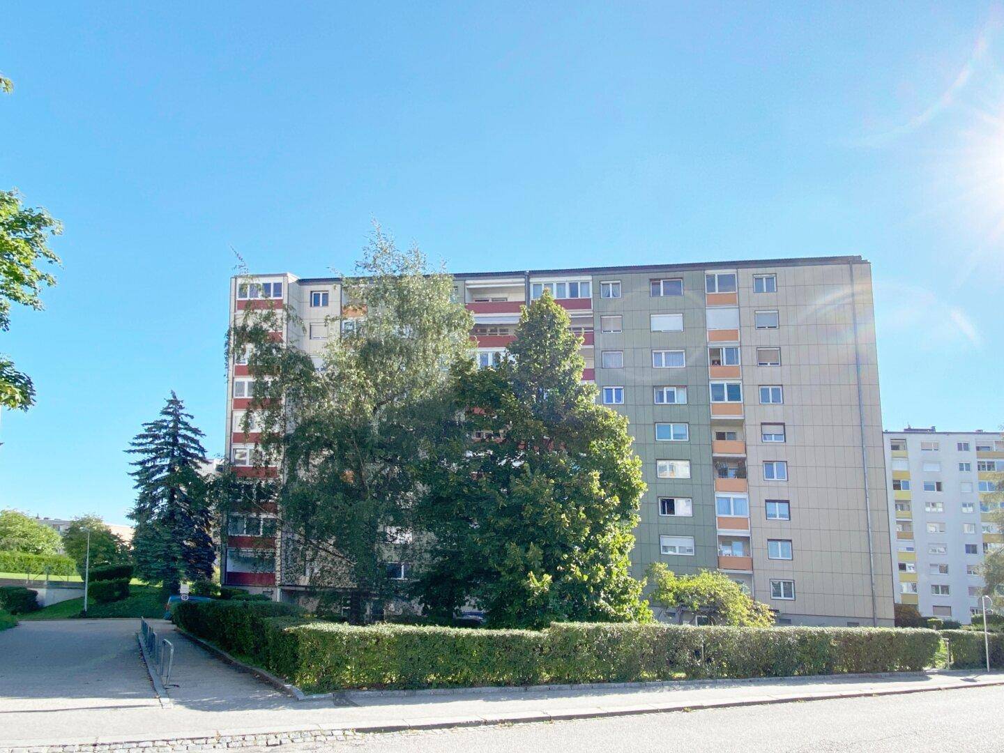 Mehrstöckiges Wohngebäude mit Balkonen und umgebender Grünfläche unter blauem Himmel.