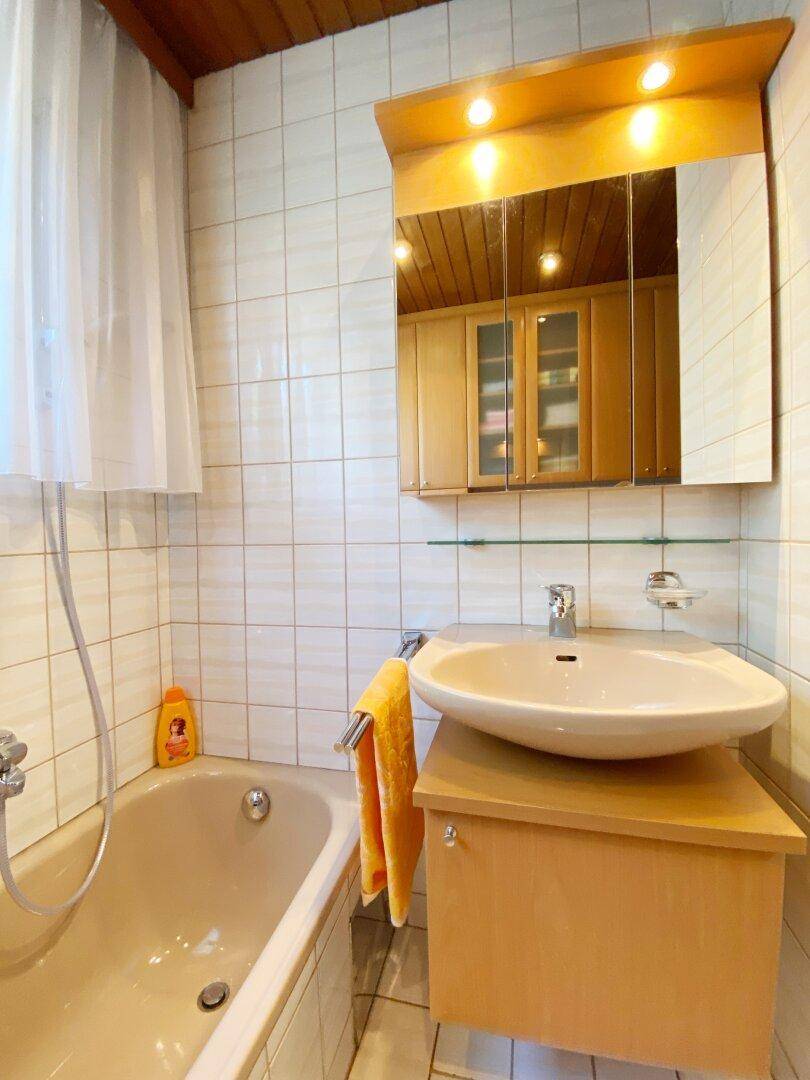 Badezimmer mit Badewanne, Waschbeckenunterschrank, Spiegelschrank und hellen Fliesen.