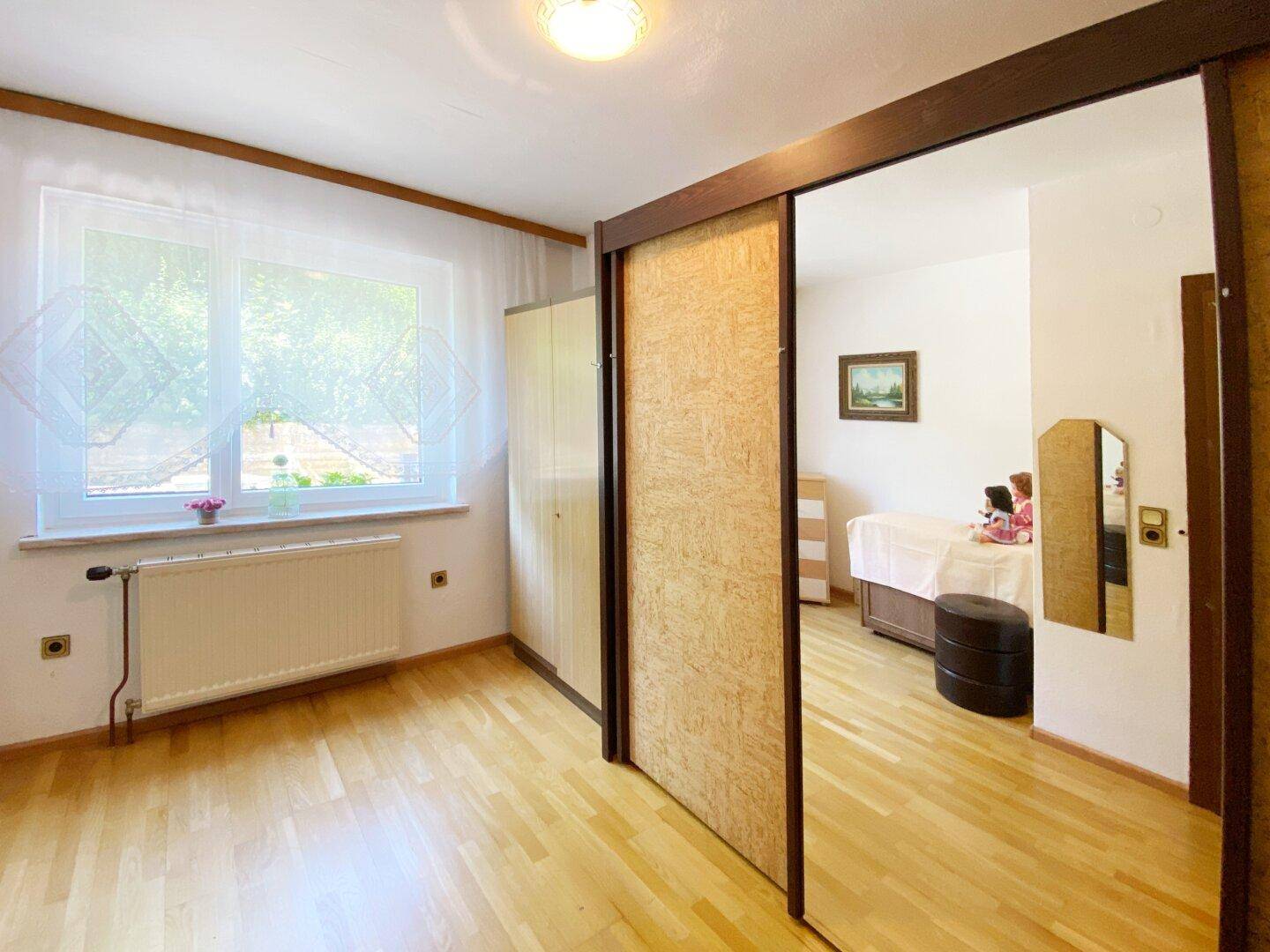 Helles Zimmer mit Holzfußboden, großem Fenster und Schiebetür zu einem weiteren Raum.