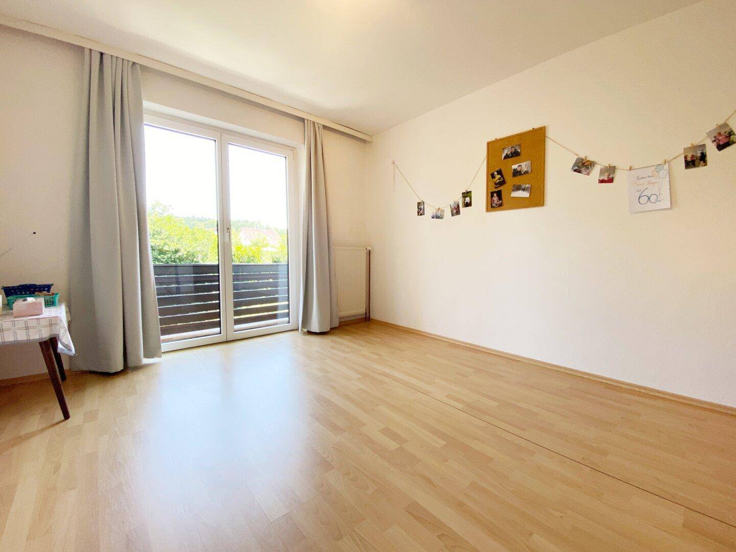 Leeres Zimmer mit Holzfußboden, Zugang zum Balkon und hellen Vorhängen.