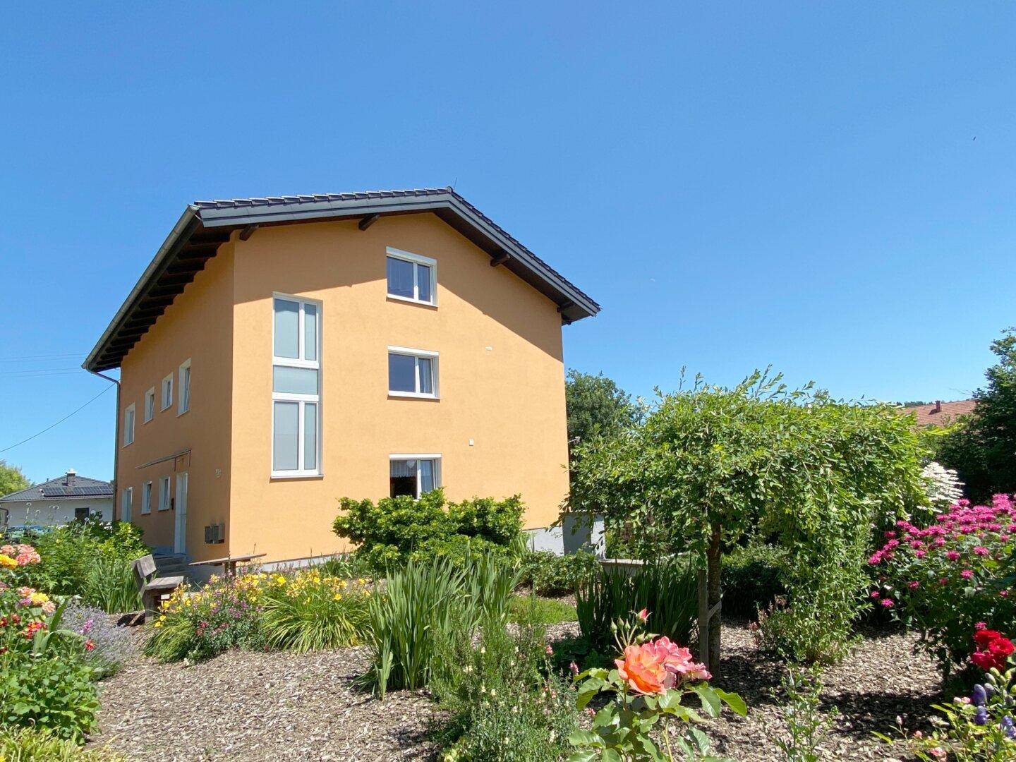 Gebäudeseite mit gepflegtem Garten, bunten Blumenbeeten und verschiedenen Bäumen unter strahlendem Himmel.