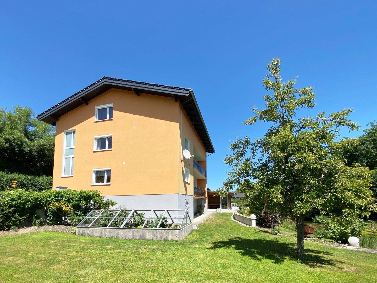 Seitenansicht des Gebäudes mit Garten, Rasenfläche, Bäumen und einem kleinen Gewächshaus.