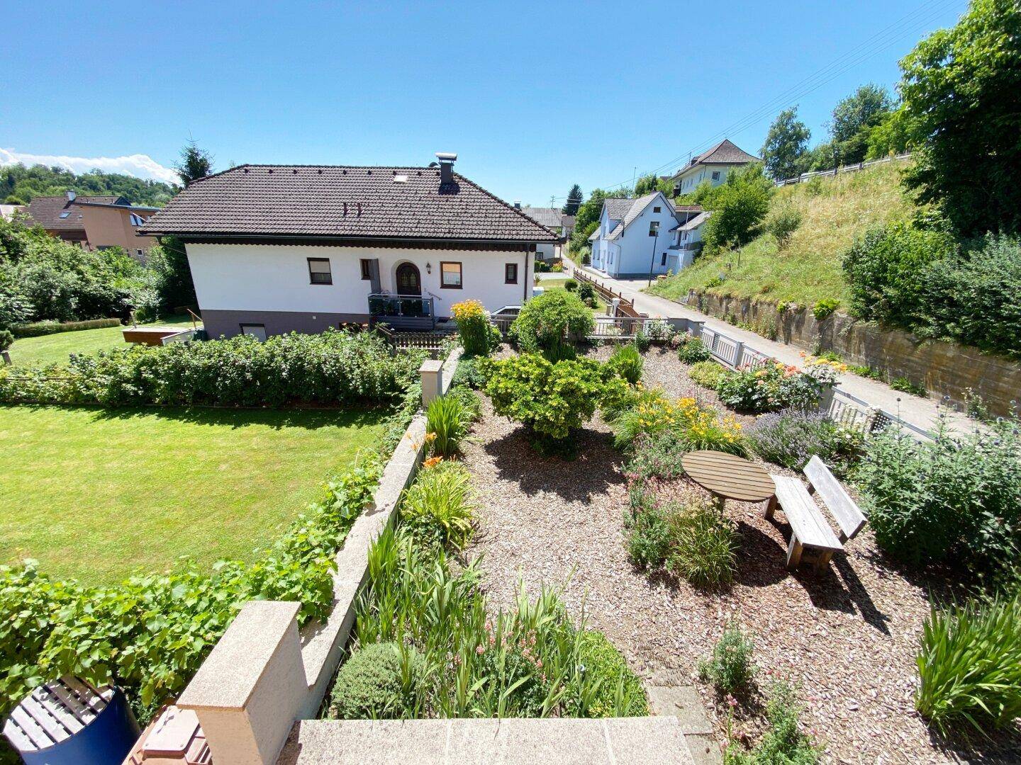 Terrassierter Garten mit vielfältiger Bepflanzung, Sitzgelegenheit und Blick auf das Wohnhaus.