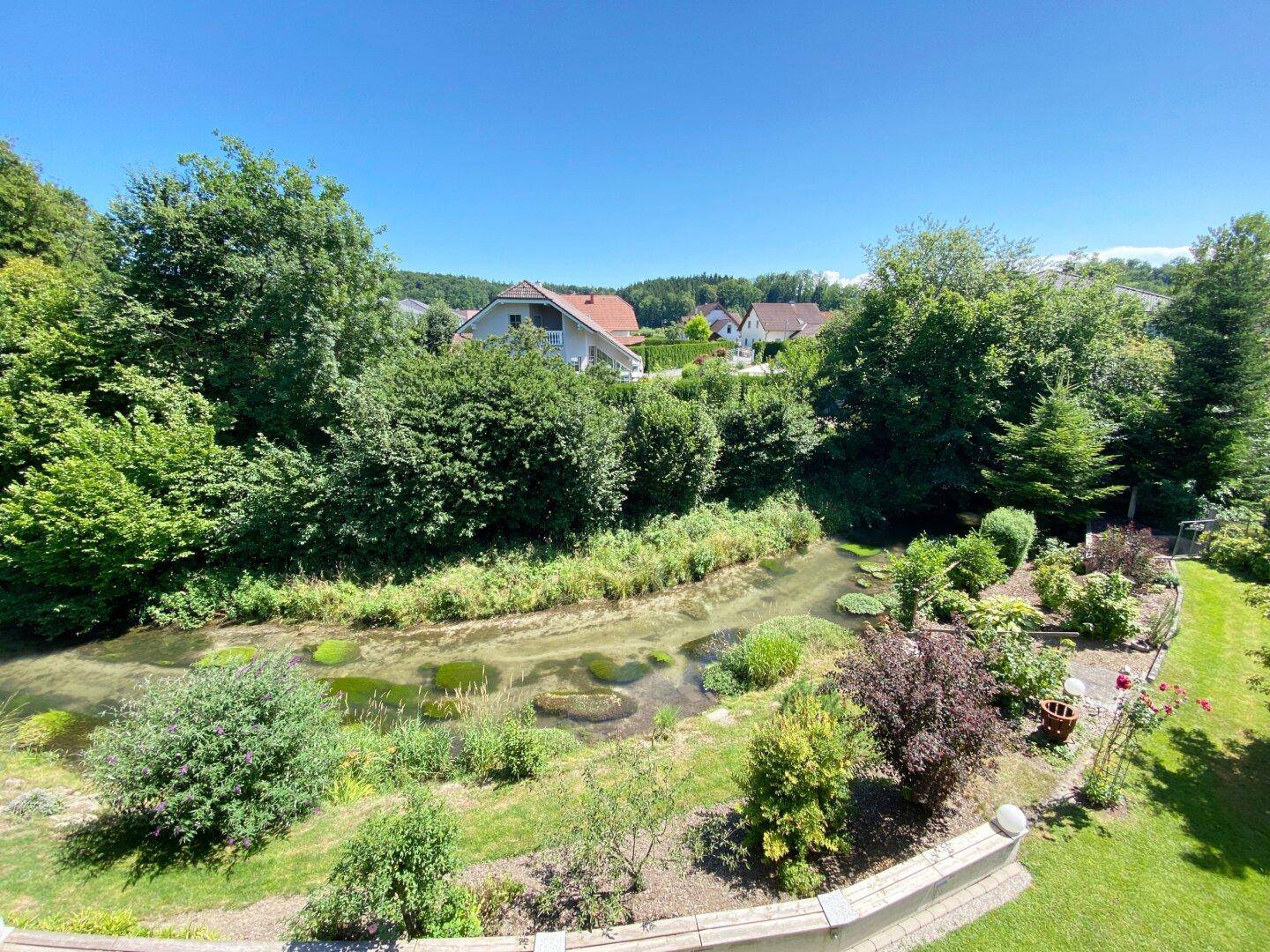Panoramablick über den Garten mit Bachlauf und den umliegenden Häusern in der Ferne.