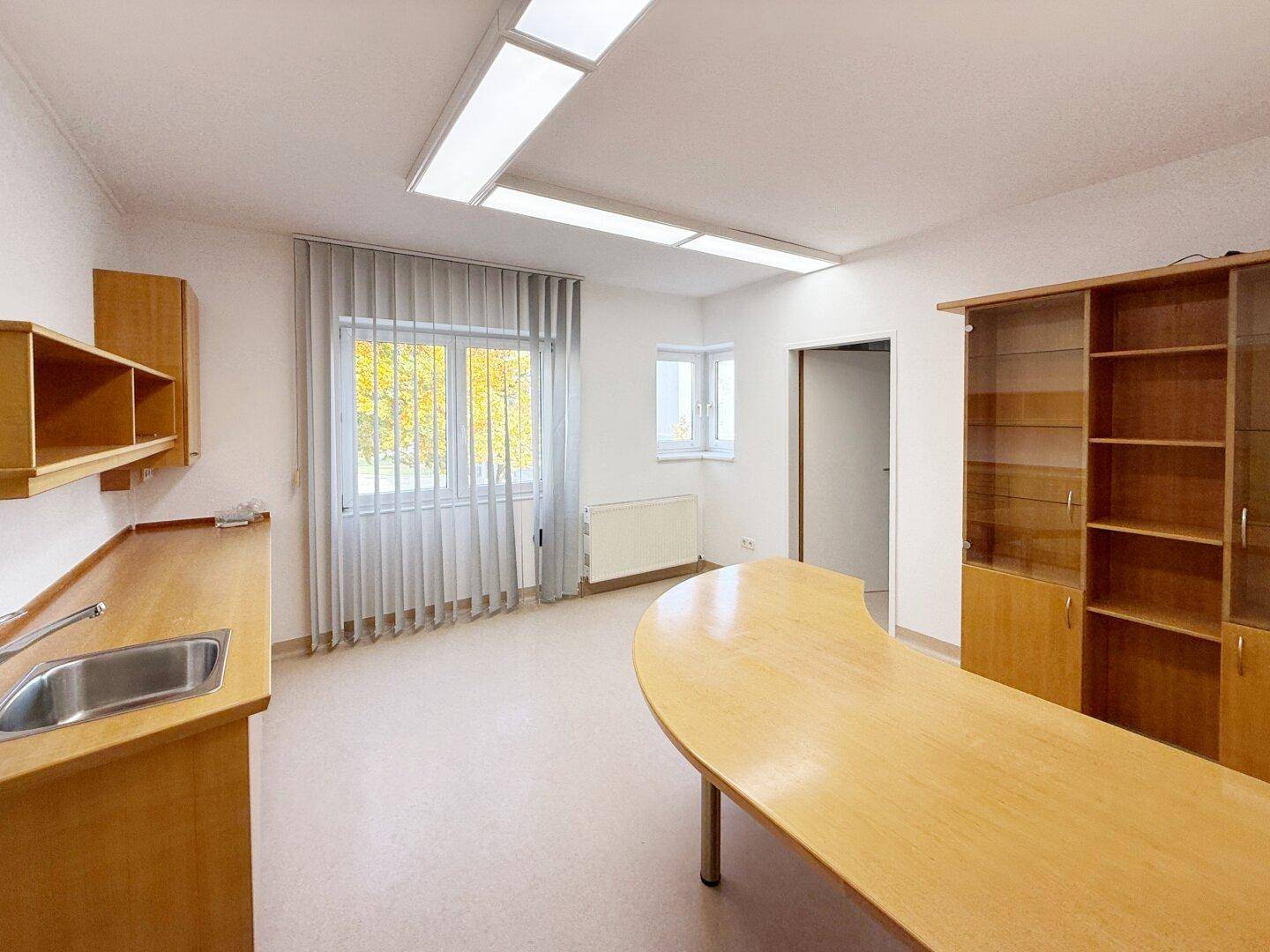 Helles Büro mit großem Schreibtisch, integrierter Teeküche und Fenstern mit Jalousien, bietet vielseitige Nutzungsmöglichkeiten.