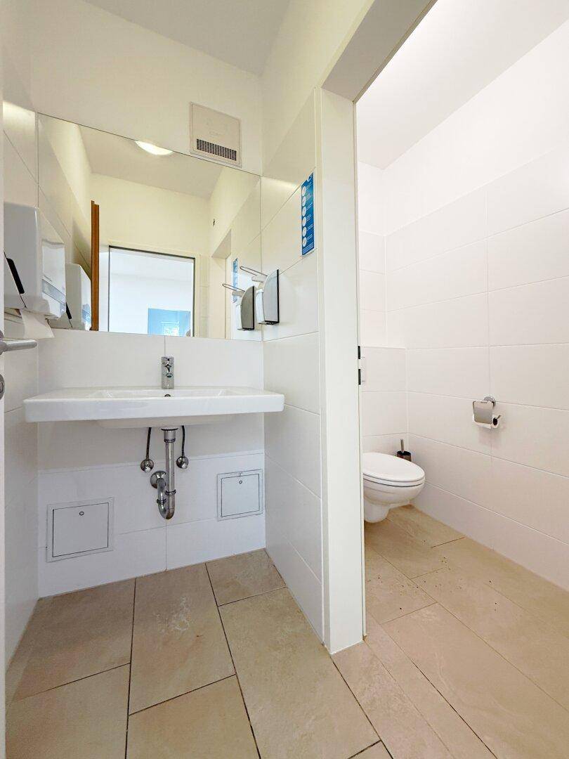 Heller Waschraum mit großem Spiegel, breitem Waschbecken und WC im Hintergrund, ideal für gewerbliche Nutzung.