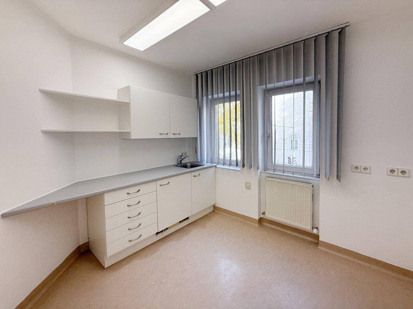 Heller Büroraum mit weißer Küchenzeile, Spülbecken und Fensterfront mit Jalousien, bietet eine praktische Arbeitsumgebung.
