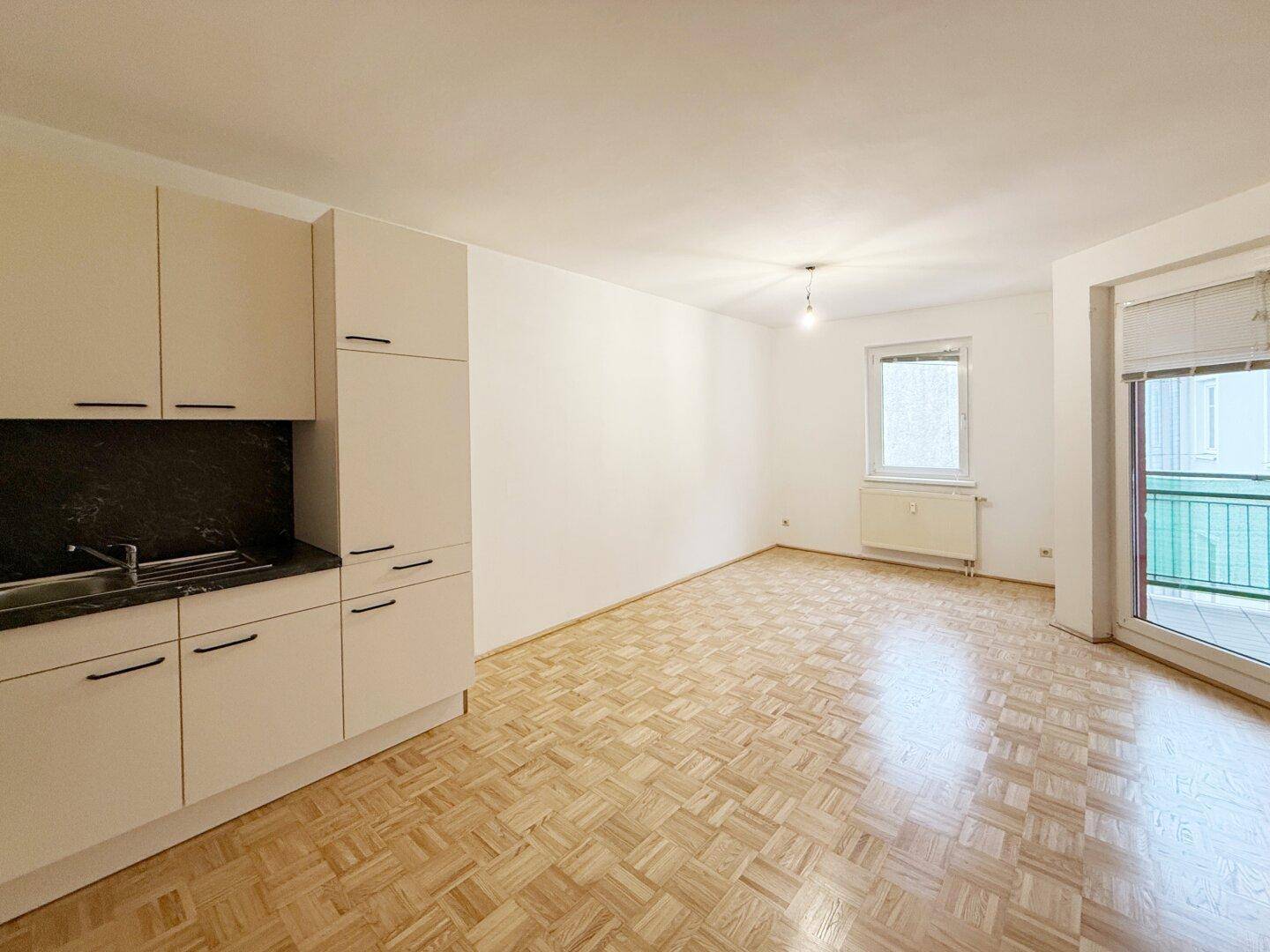 Heller Wohnbereich mit Küchenzeile, Parkettboden und Zugang zu einem Balkon.