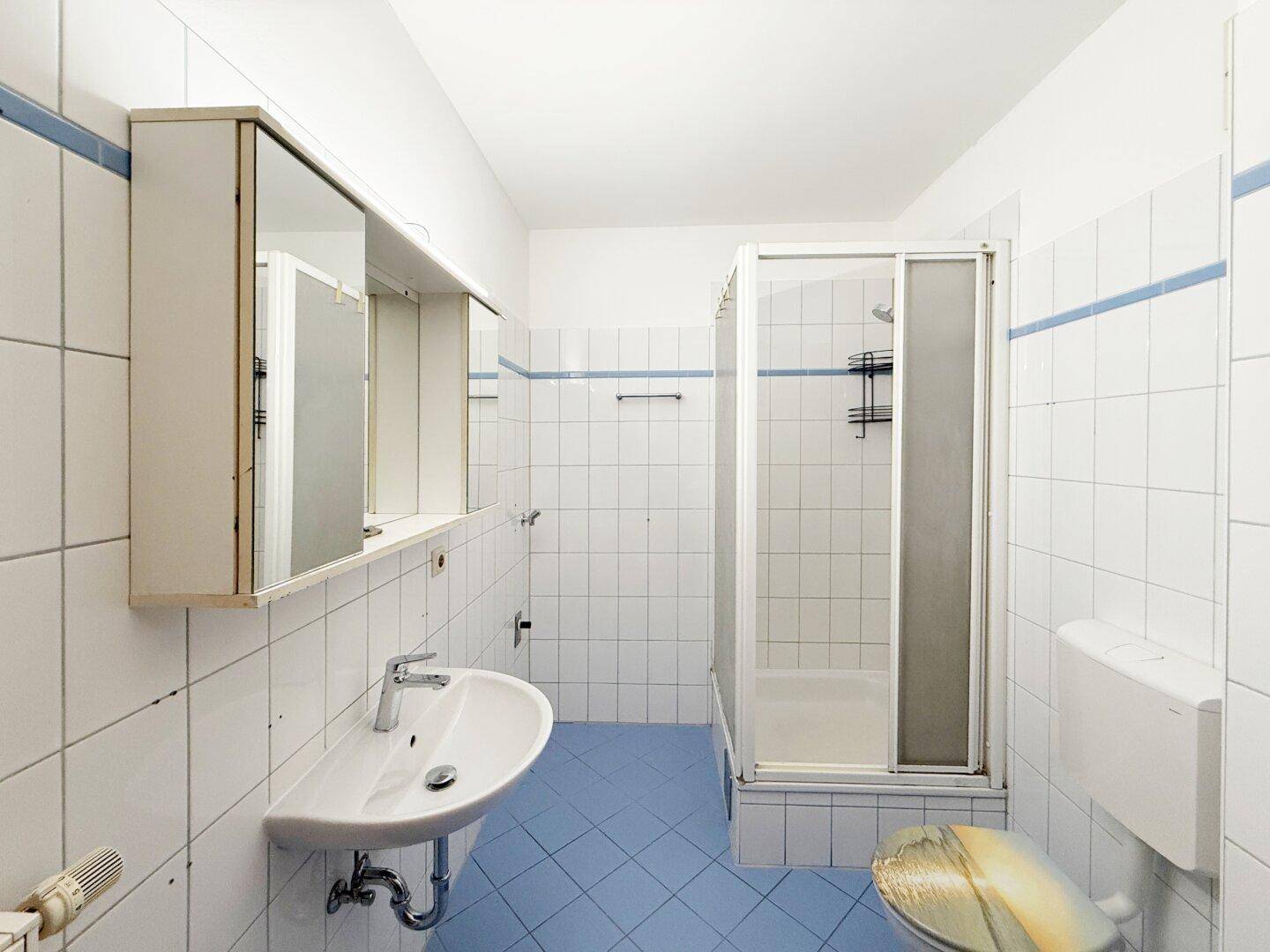 Funktionales Badezimmer mit weißen Fliesen, blauer Bodenfliese und Duschkabine.
