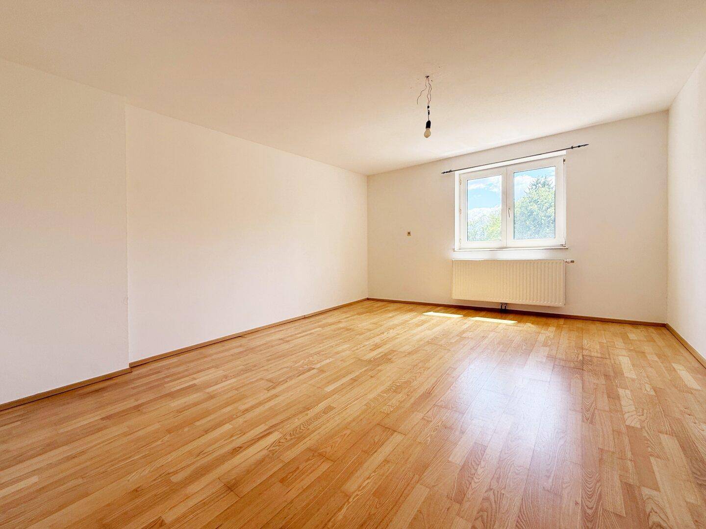 Geräumiger Wohnbereich mit hellem Parkettboden und Fenster mit Blick ins Freie.