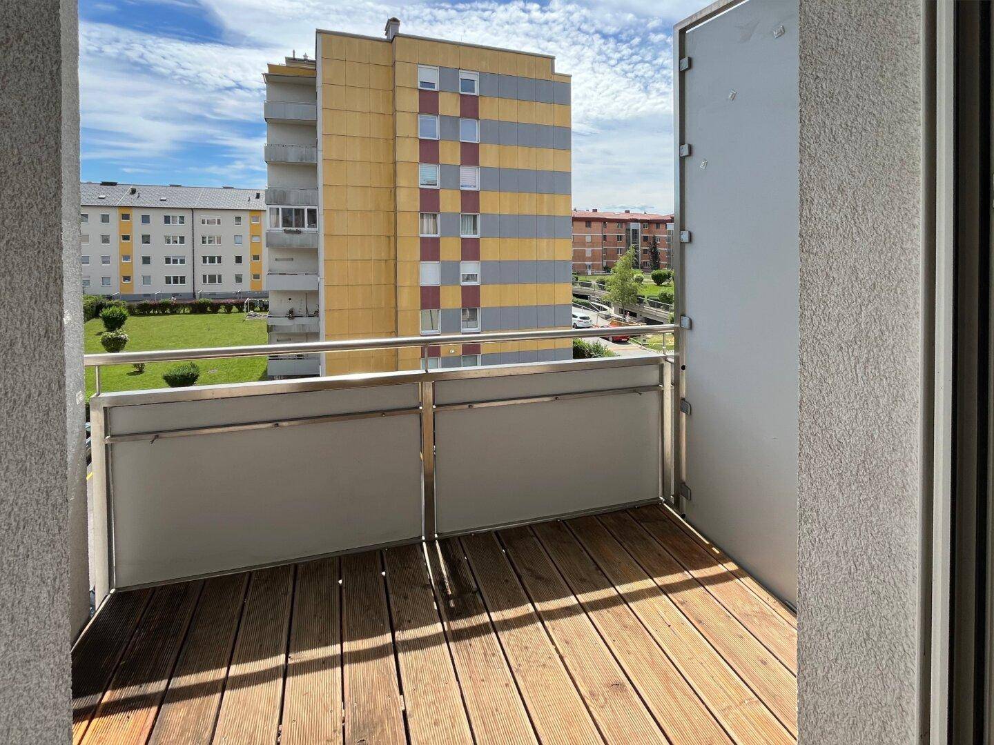 Balkon mit Holzboden und Blick auf umliegende Wohngebäude und den Himmel.