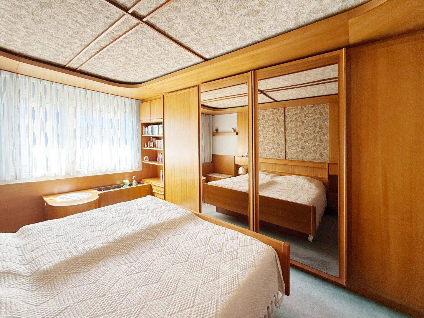 Schlafzimmer mit Spiegelschränken, Holzvertäfelung und Fenster mit Vorhängen.