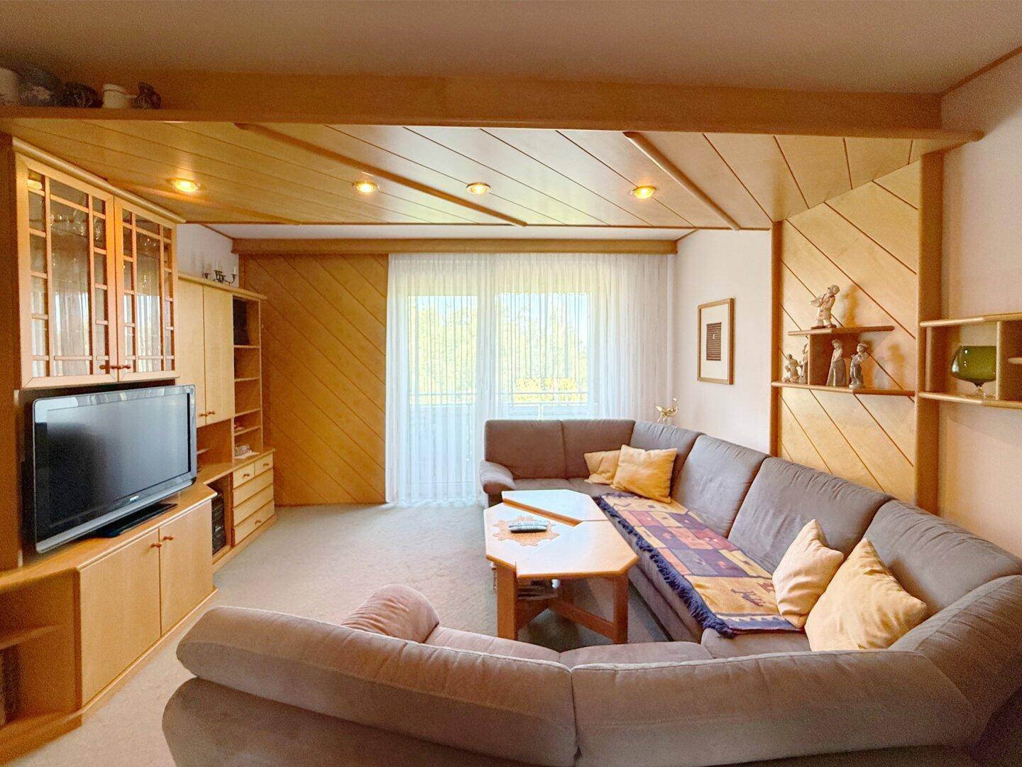 Wohnzimmer mit Fernsehmöbeln, Holzvertäfelung und Blick auf den Balkon durch Vorhänge.