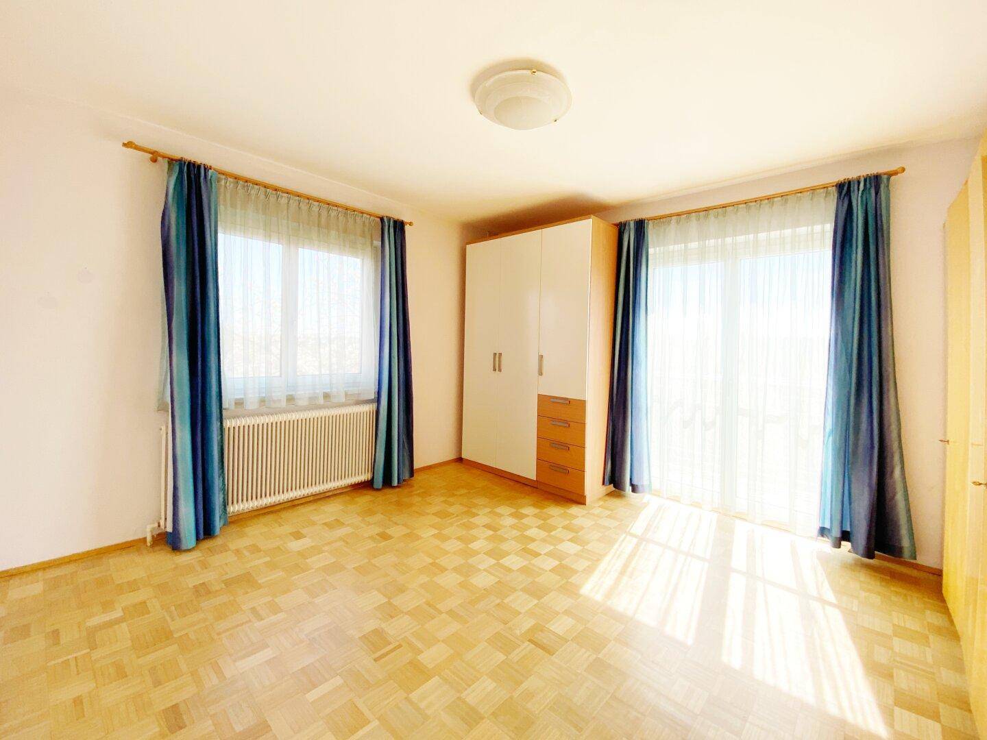 Helles Schlafzimmer mit Parkettboden, Kleiderschrank und blauen Vorhängen an den Fenstern.