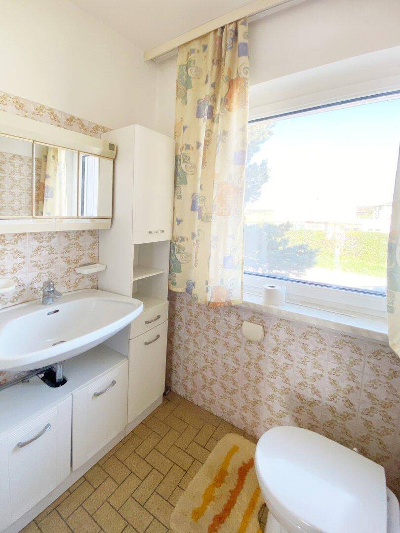 Badezimmer mit WC, Waschbecken, Spiegelschrank und gemusterten Fliesen im 80er-Jahre-Stil.