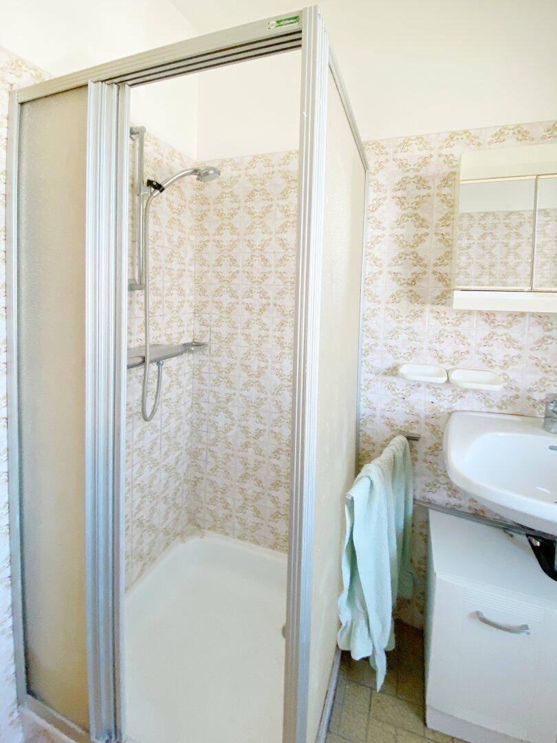 Funktionales Badezimmer mit Duschkabine, Waschbecken und gemusterten Fliesen im 80er-Jahre-Stil.