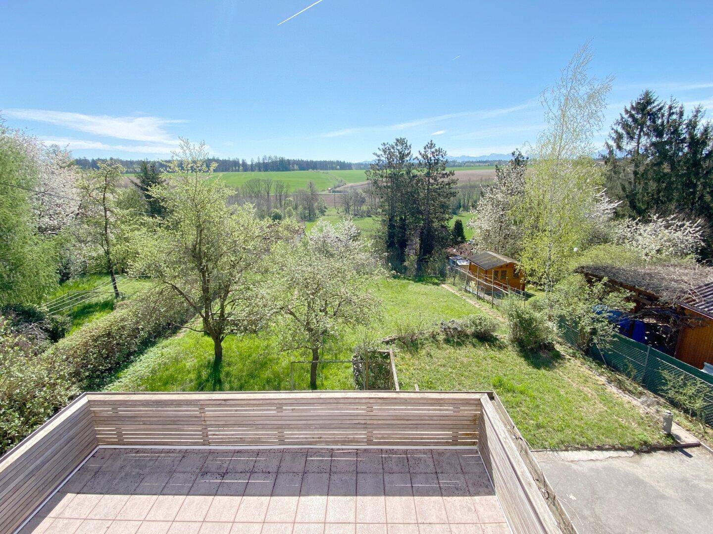 Geräumige Terrasse mit Holzelementen und weitem Blick über grüne Felder und Bäume unter blauem Himmel.