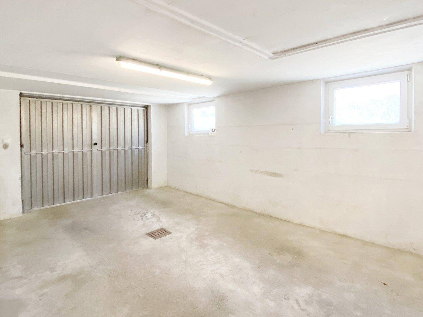 Geräumige Garage mit Betonboden, zwei Fenstern und einem großen Metalltor.