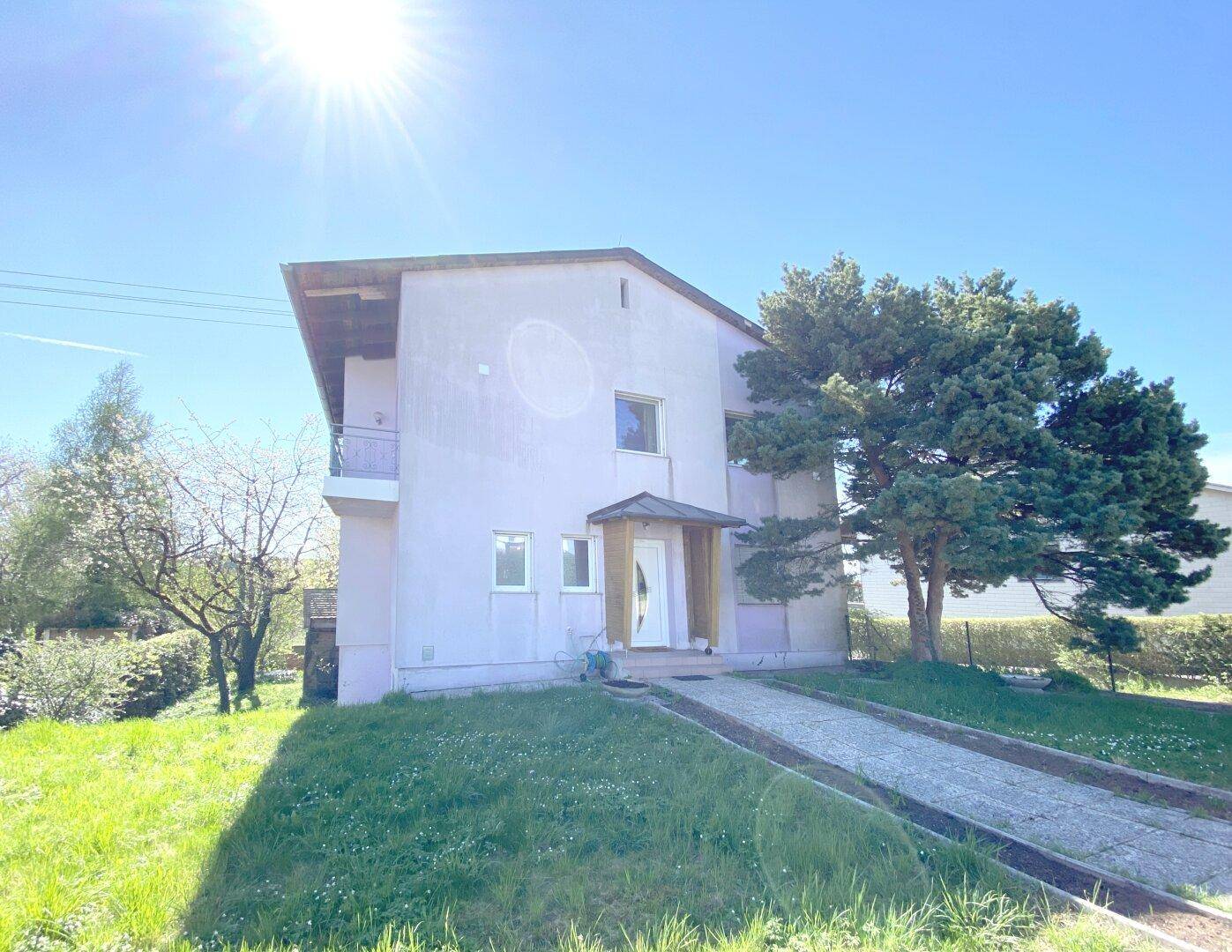 Einfamilienhaus mit heller Fassade, umgeben von Grünflächen und einem sonnigen Vorgarten mit Auffahrt.