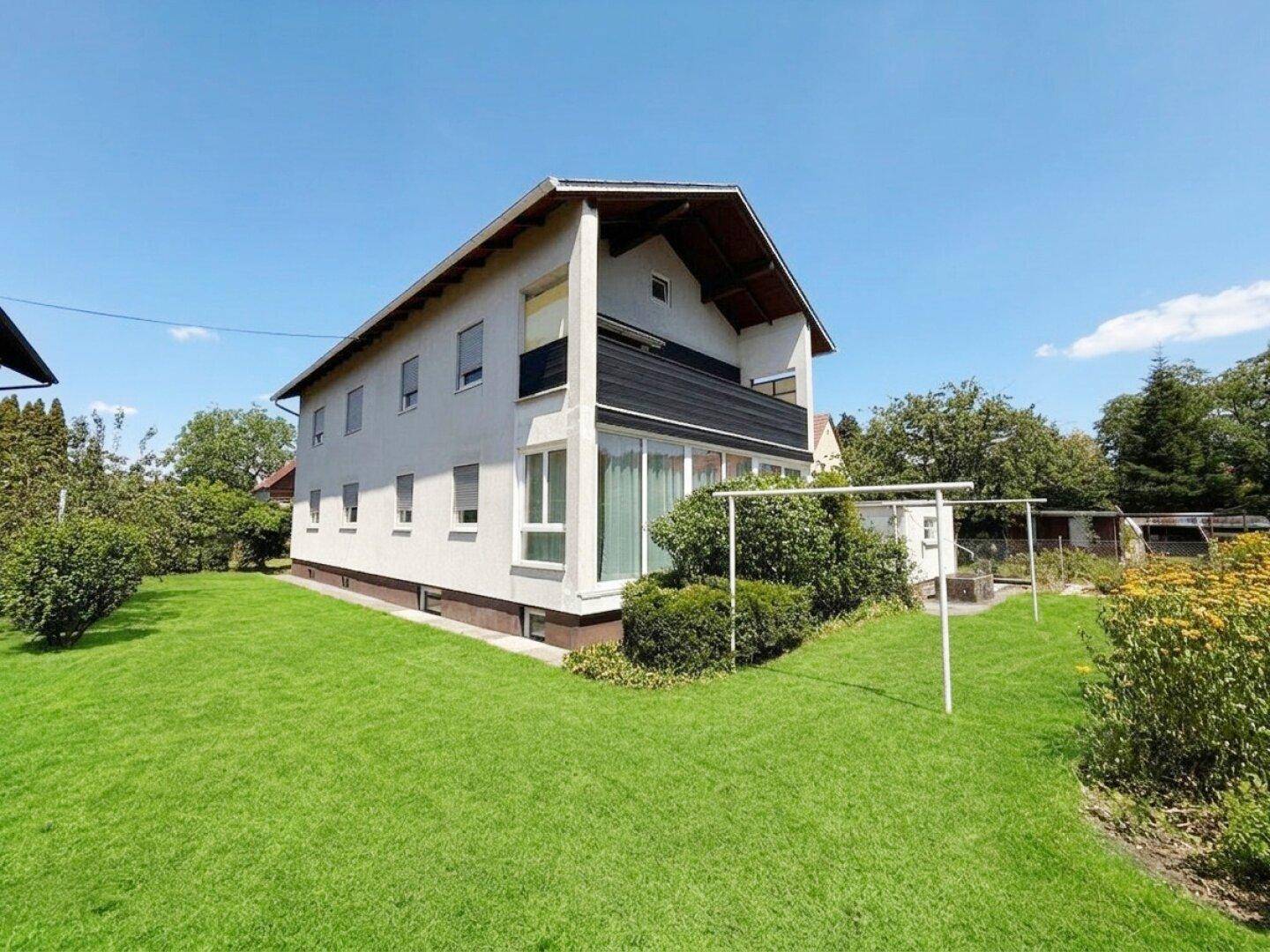 Hausfassade mit großem Gartenbereich und Rasenfläche unter blauem Himmel.