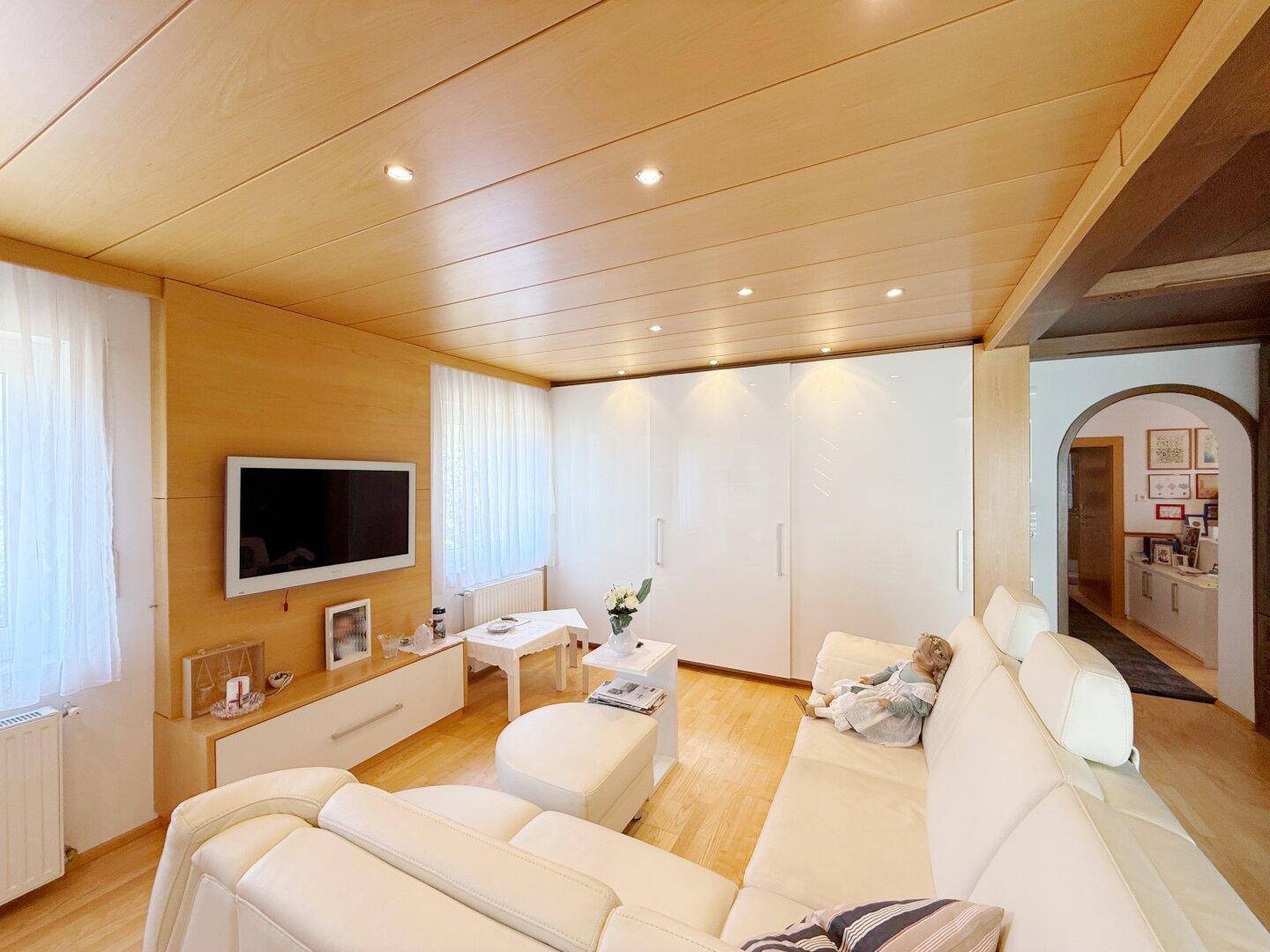 Modernes Wohnzimmer mit heller Ledercouch, Holzdecke und integrierter Beleuchtung.