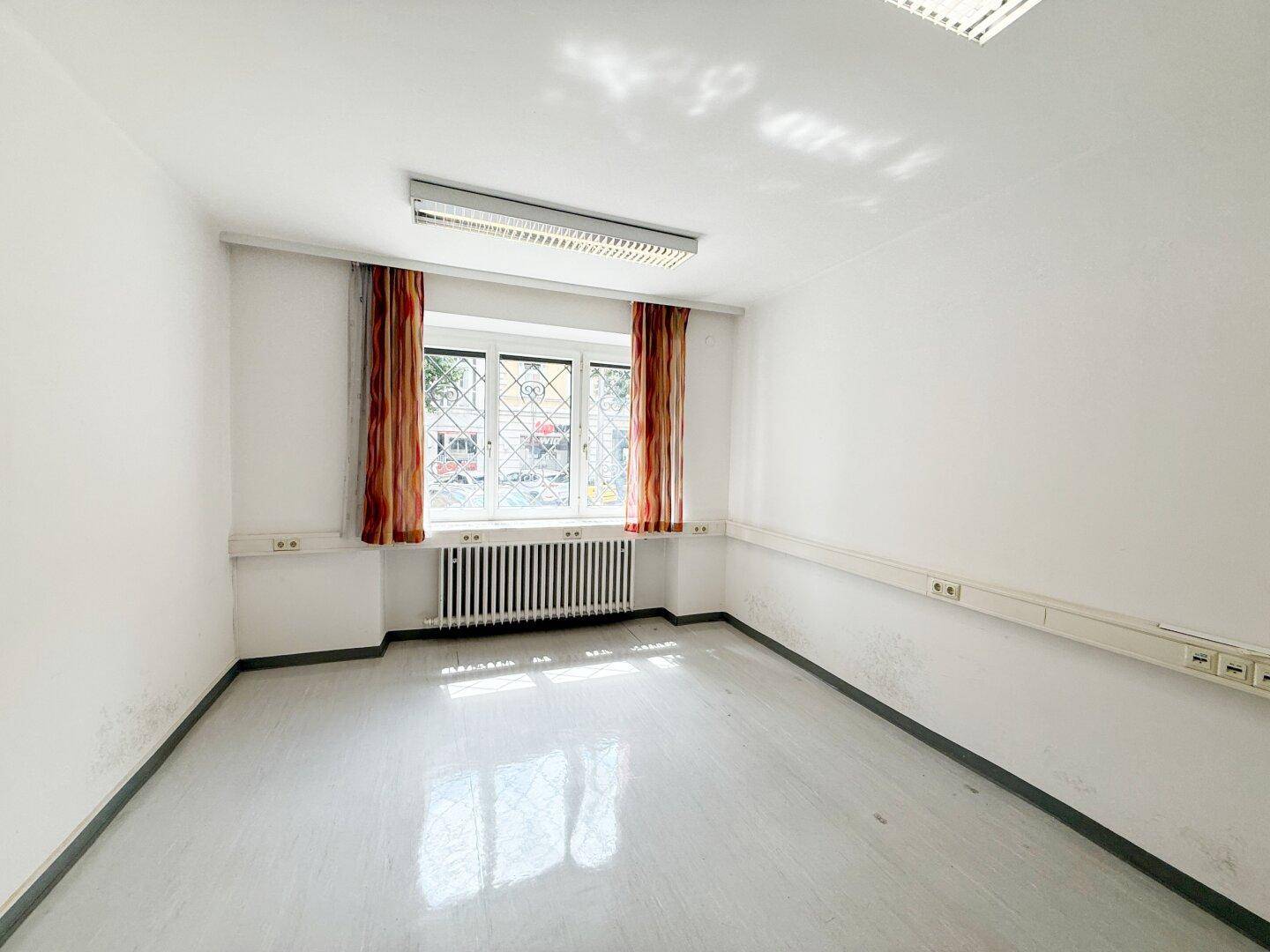 Leerer Büroraum mit großem Fenster, Heizkörper und schlichten Vorhängen.