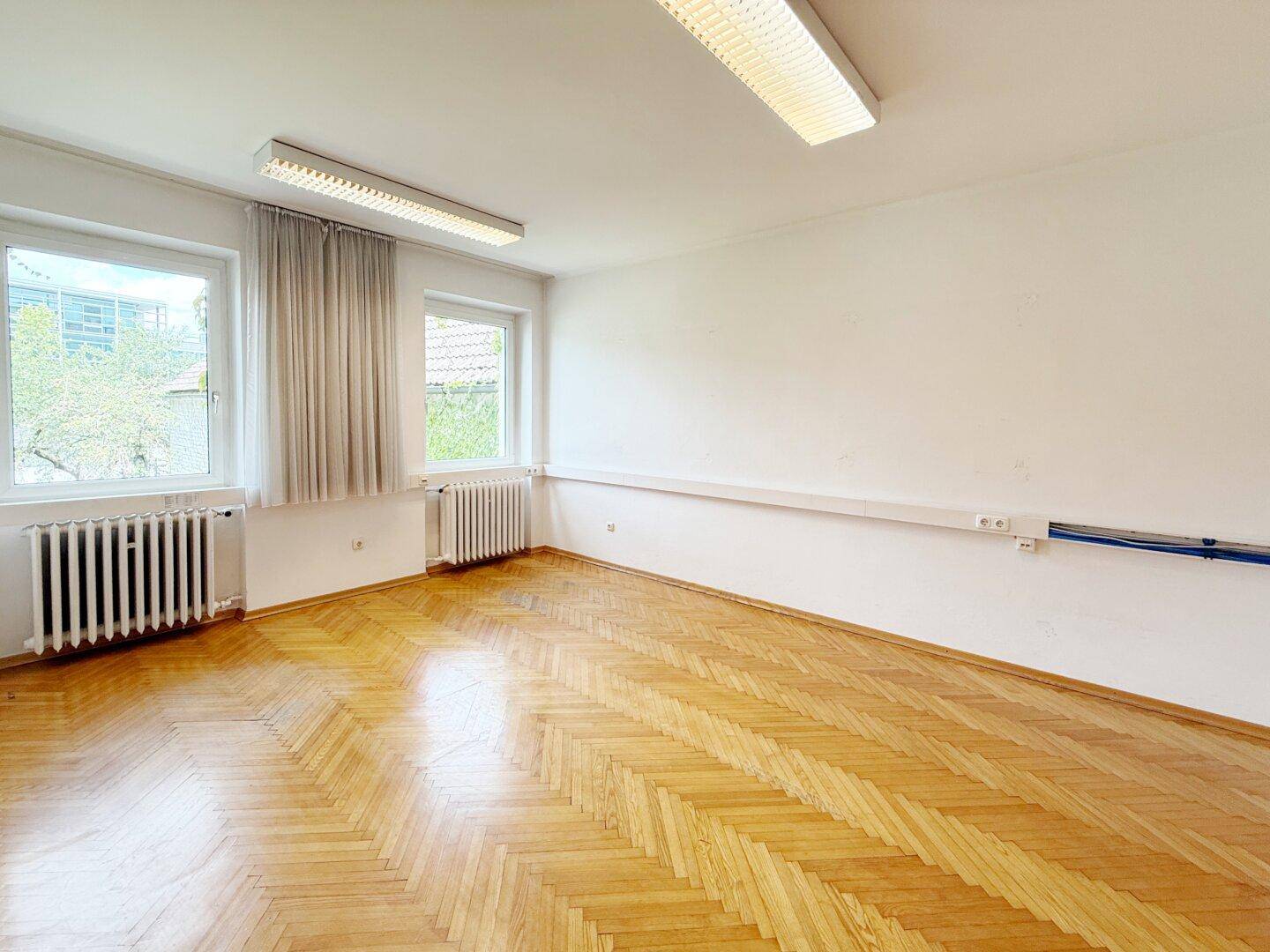 Geräumiger Büroraum mit Fischgrätparkett, zwei Fenstern und Heizkörpern.