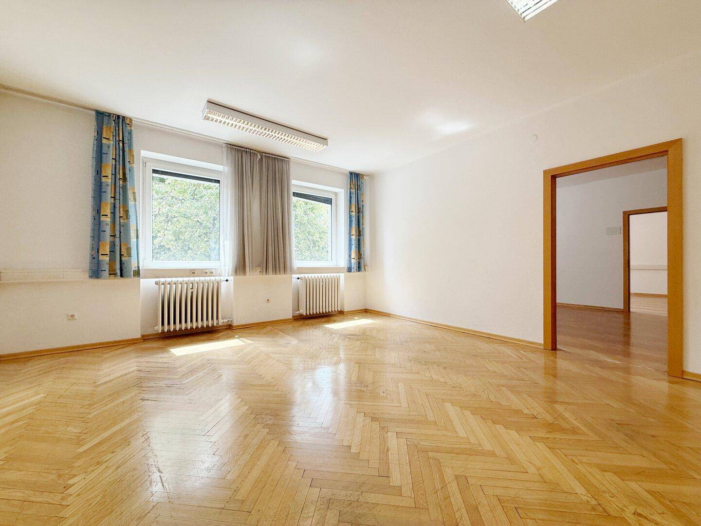 Heller Büroraum mit klassischem Fischgrätparkett, Fenstern und Zugang zu weiterem Raum.