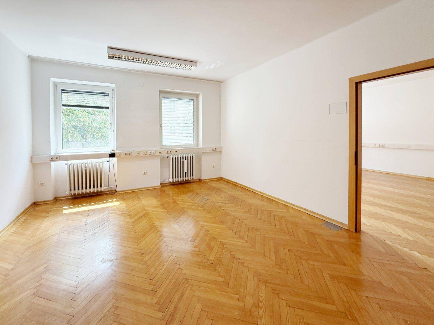 Büroraum mit Fischgrätparkett, Fenstern mit Jalousien und Zugang zu einem weiteren Bereich.