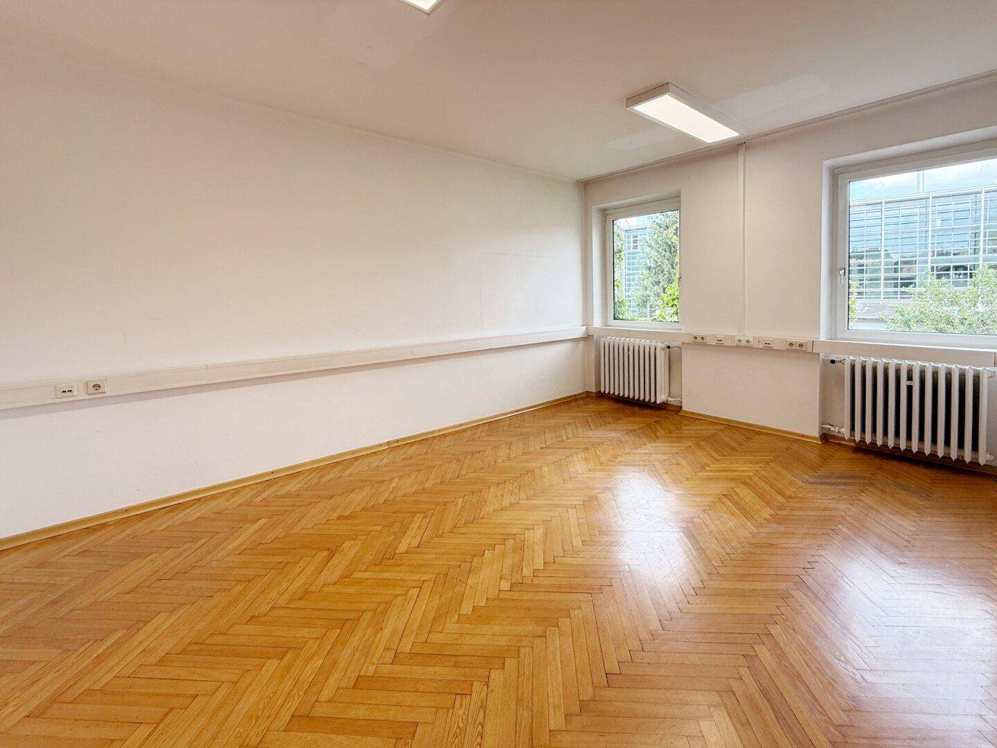 Heller Büroraum mit Fischgrätparkett, zwei Fenstern und Blick ins Grüne.