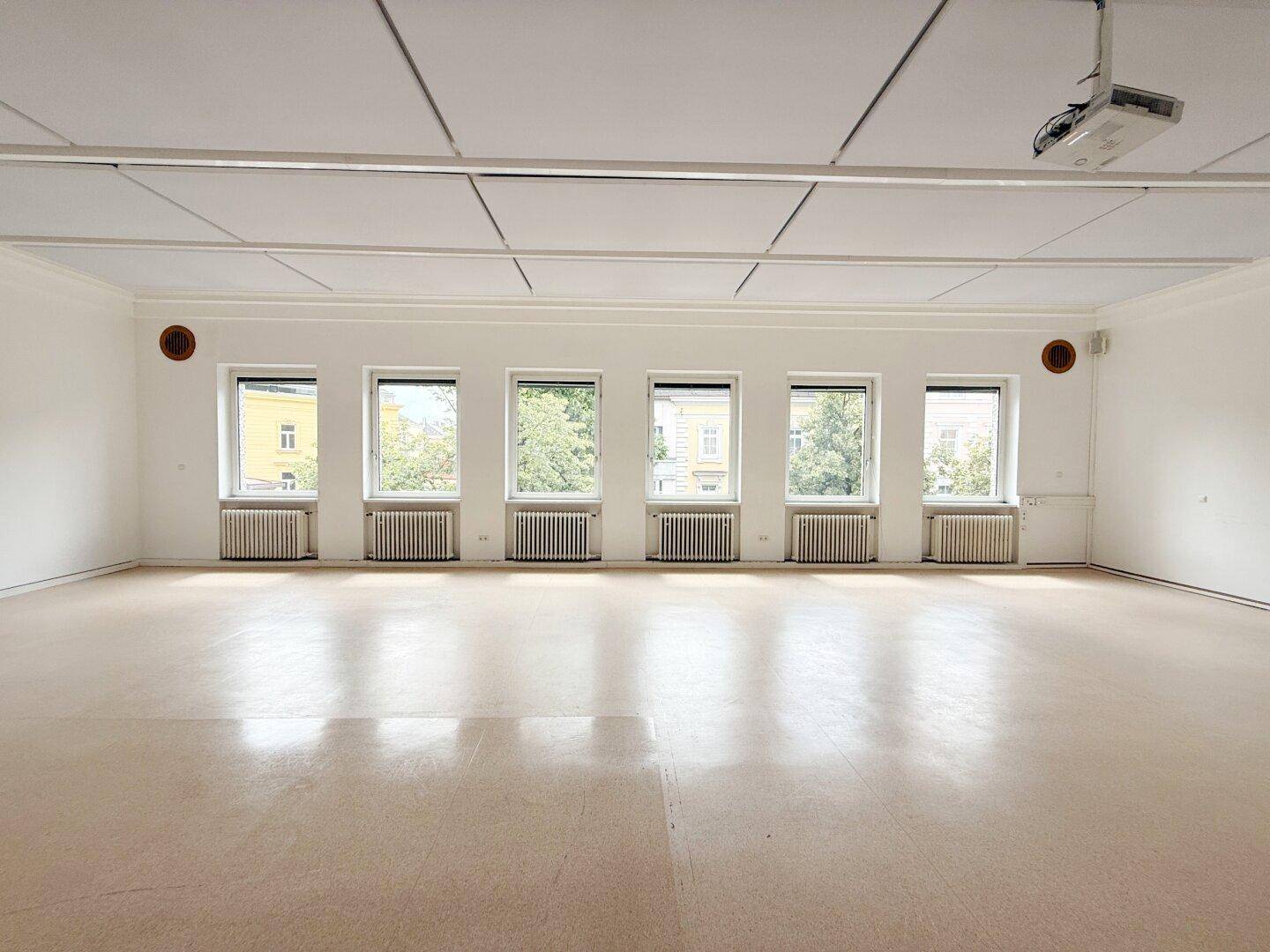 Großer, heller Raum mit vielen Fenstern, abgehängter Decke und Beamer.