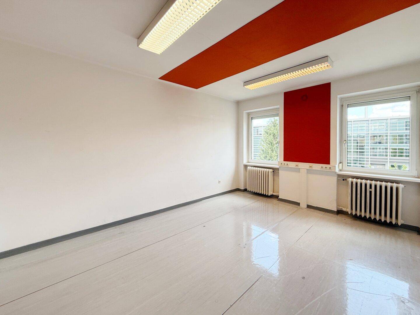 Büroraum mit roter Akzentwand, zwei Fenstern und glänzendem Boden.
