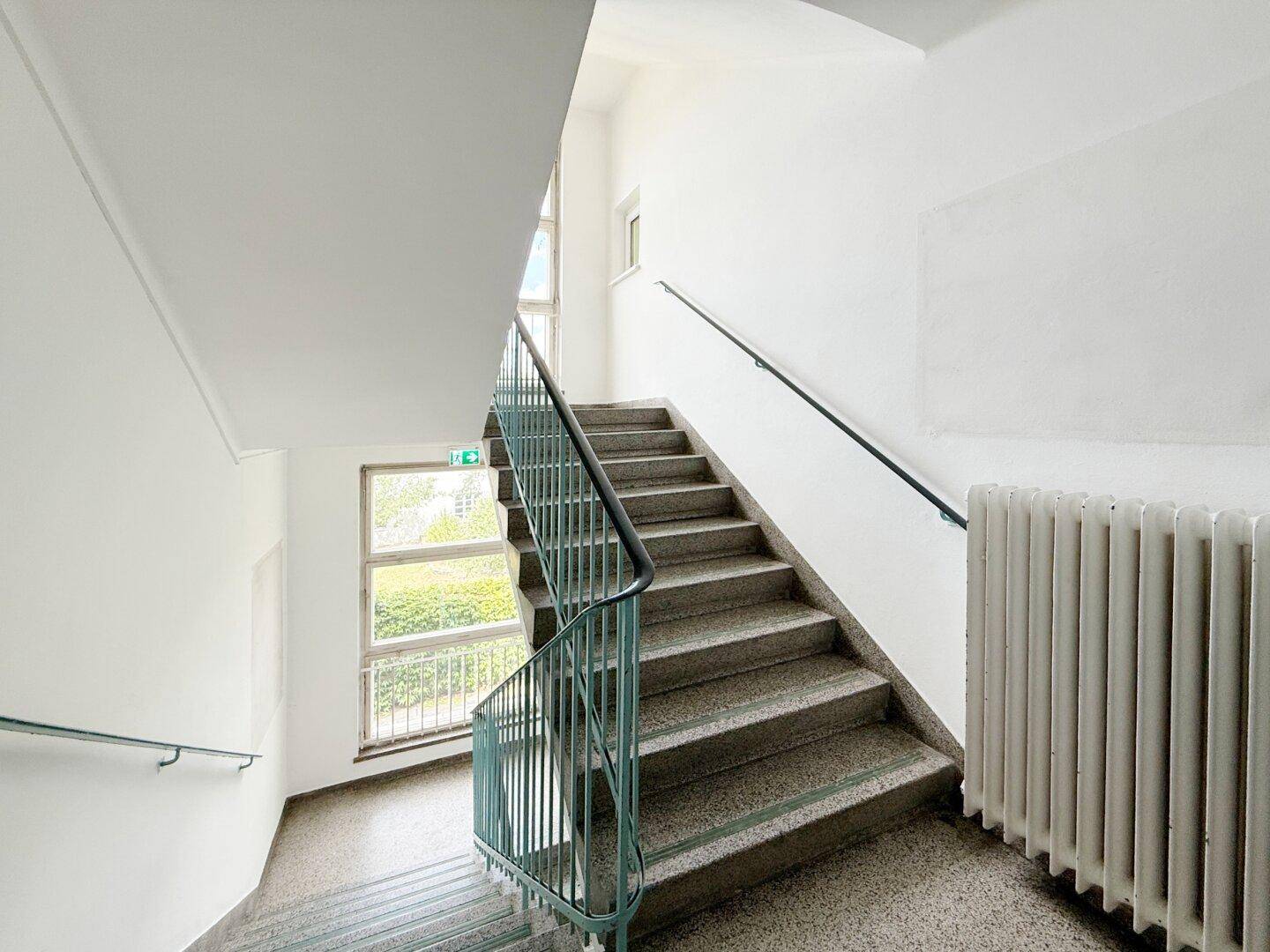 Helles Treppenhaus mit Terrazzoboden, Geländer und Fenstern für Tageslicht.