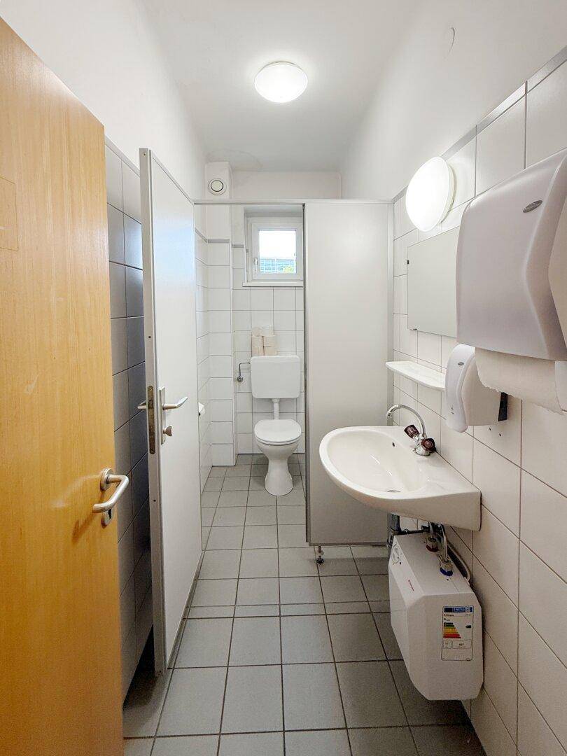 Gefliestes WC mit mehreren Kabinen, Waschbecken und kleinem Fenster.