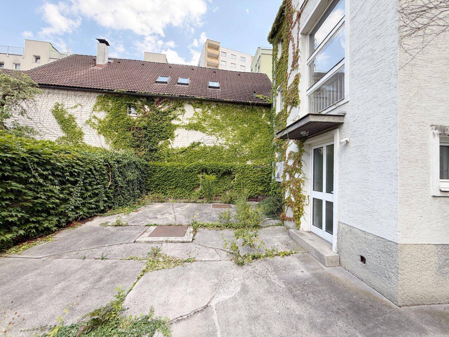 Hinterhof mit Betonfläche, umgeben von Hecken und bewachsenen Mauern.