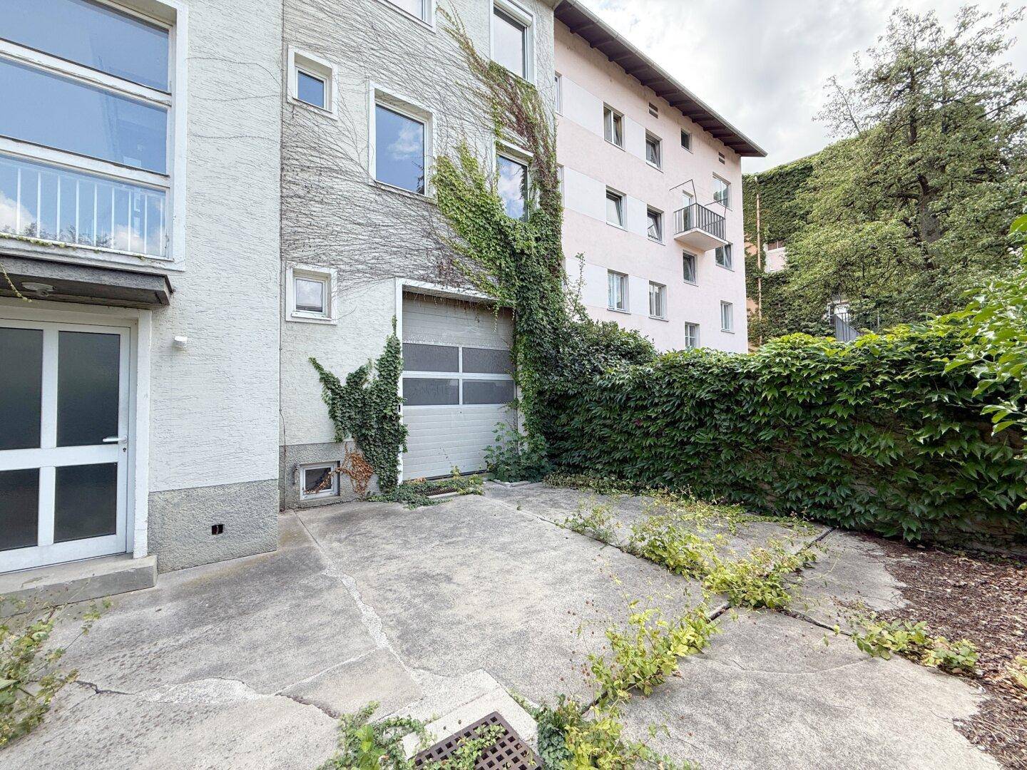 Hinterhof mit Betonfläche, Garagentor und hohen Hecken.