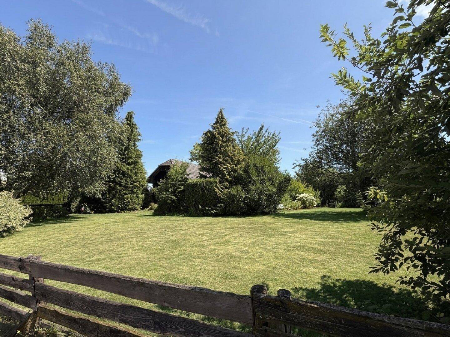 Sonniger Garten mit großer Rasenfläche, altem Baumbestand und rustikalem Holzzaun im Vordergrund.